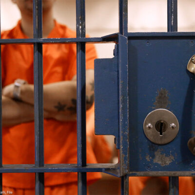 The lock and bars of a prison cell are at the forefront, while a detainee, standing in an orange jumpsuit and with his tattooed arms crossed waits in a cell with other detainees.