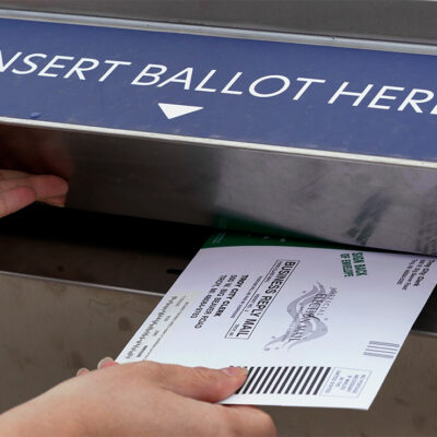 A voter holds the door of a ballot box open with her left hand while inserting her voter ballot with her right hand.