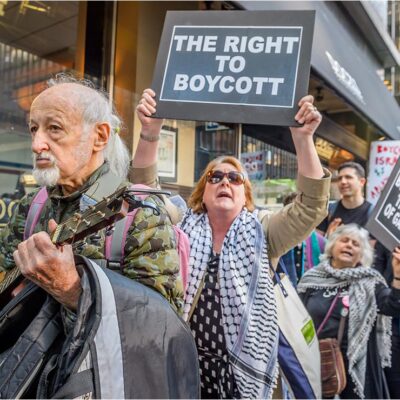 Protestors being led by a man playing a guitar, followed by women holding signs saying" THE RIGHT TO BOYCOTT", and "BOYCOTT UNTIL THE SIEGE ON GAZA ENDS".