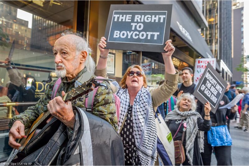 Protestors being led by a man playing a guitar, followed by women holding signs saying" THE RIGHT TO BOYCOTT", and "BOYCOTT UNTIL THE SIEGE ON GAZA ENDS".