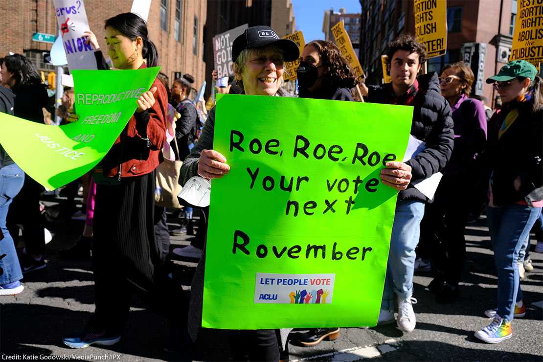 A woman holding a sign reading "Roe, Roe, Roe, Your vote next Rovember / LET PEOPLE VOTE ACLU" poses for the camera as fellow activists walk behind her.