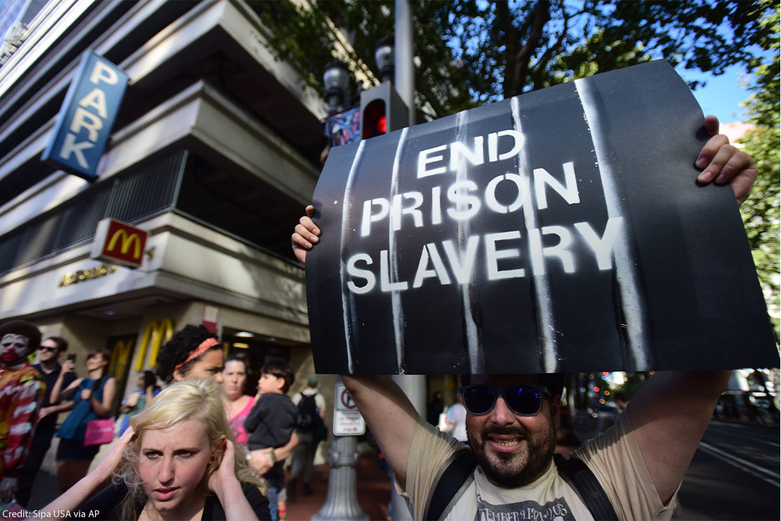 A bearded man wearing sunglasses holds up a black sign with white lettering reading "End Prison Slavery" at a protest in downtown Portland, Ore., during a nationwide day of action against prison slavery on the 45th anniversary of the Attica Uprising.