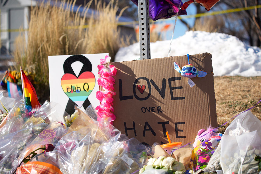 A white sign with a black ribbon in the background and a rainbow heart with the words "Club Q" in the foreground along with a cardboard sign reading "LOVE over HATE" in large letters sit above bouquets of flowers on a corner near Club Q.
