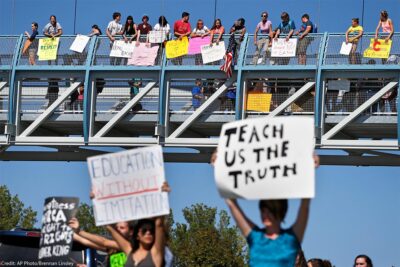 In the foreground (at an intersection,) two protesters carry signs with one reading "EDUCATION WITHOUT LIMITATION" and the other "TEACH US THE TRUTH", while in the background, other student demonstrators line an overpass protesting a proposal to emphasize patriotism and downplay civil unrest in the teaching of U.S. history.