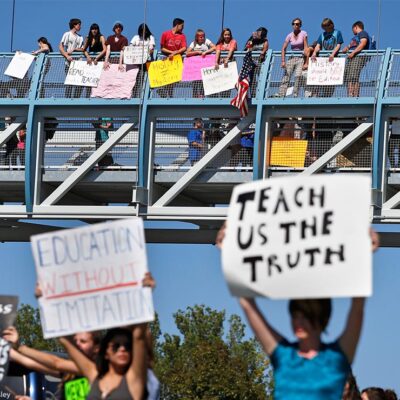 In the foreground (at an intersection,) two protesters carry signs with one reading "EDUCATION WITHOUT LIMITATION" and the other "TEACH US THE TRUTH", while in the background, other student demonstrators line an overpass protesting a proposal to emphasize patriotism and downplay civil unrest in the teaching of U.S. history.