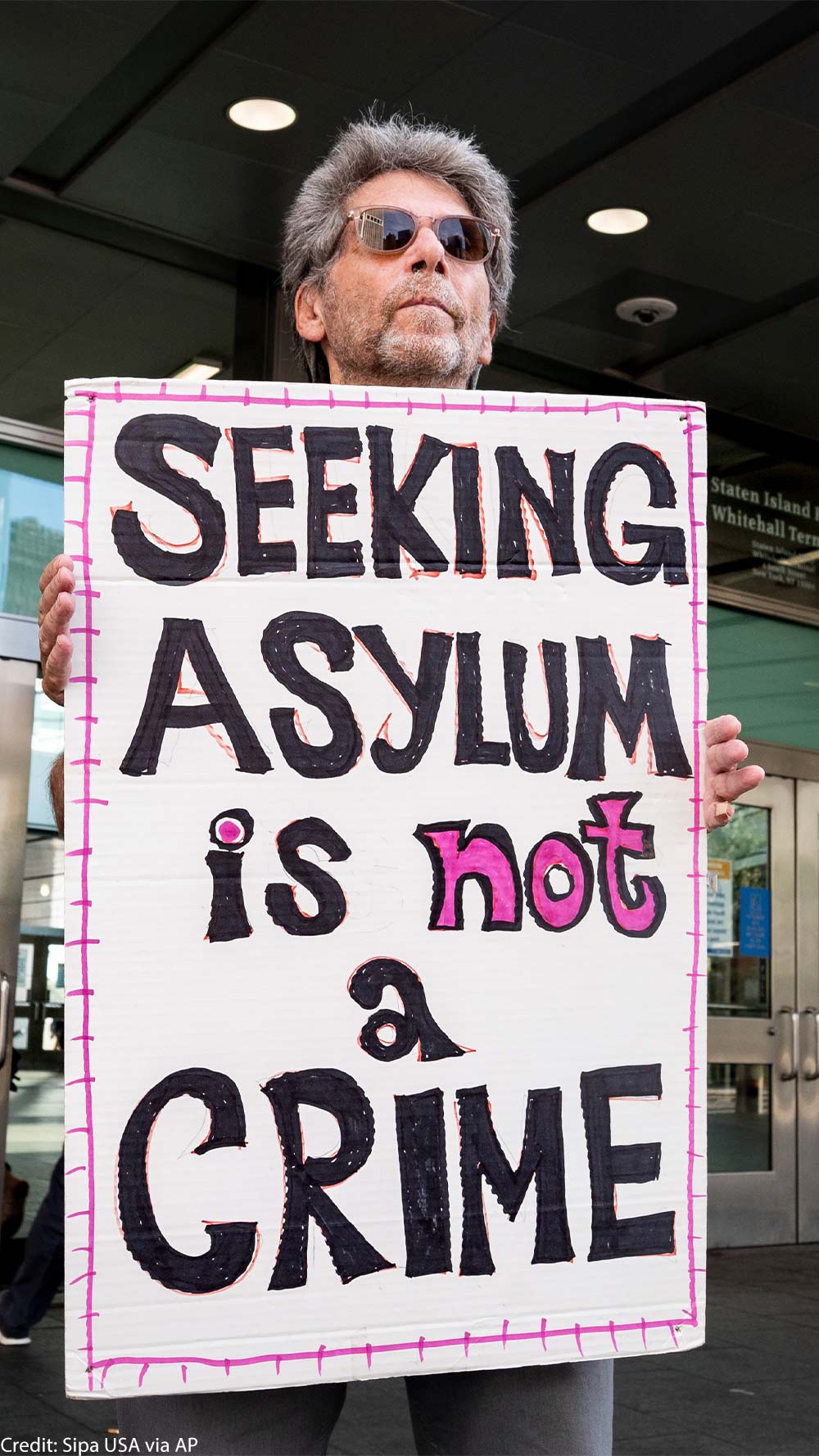 An individual in sunglasses holding a sign that says "Seeking Asylum is not a Crime."
