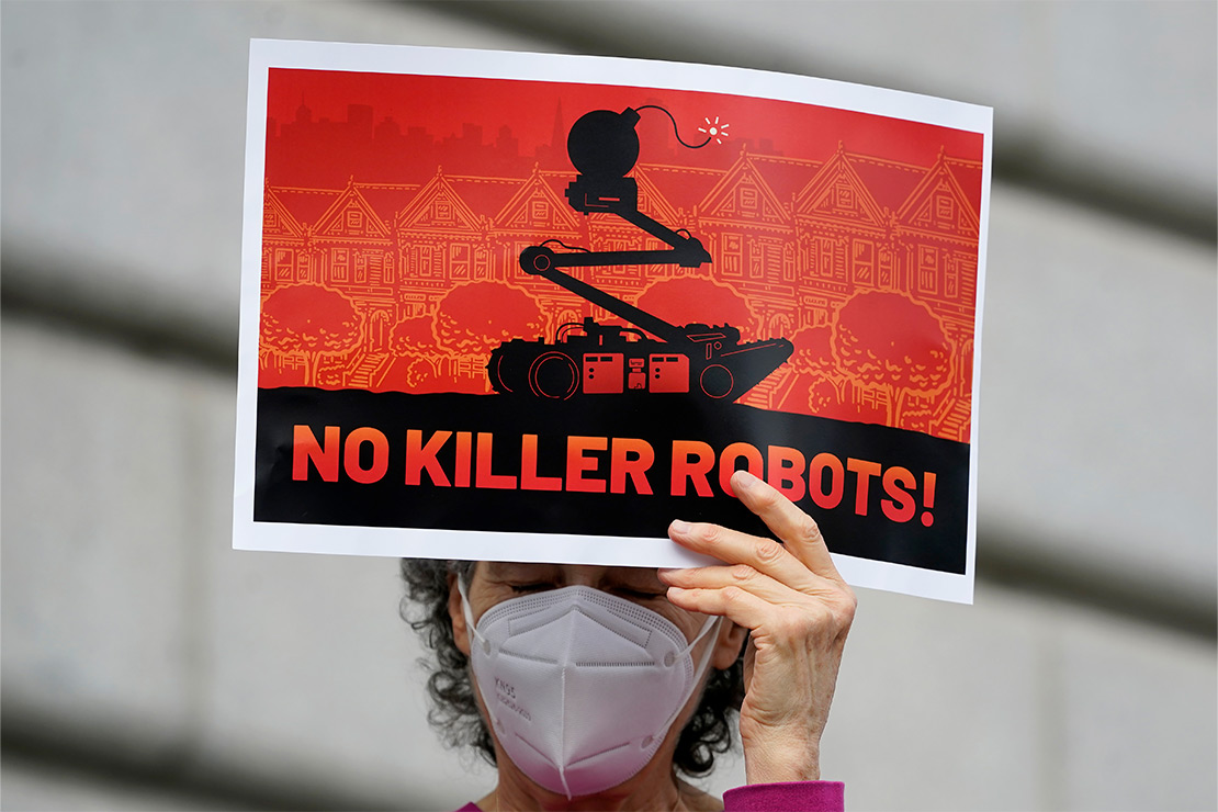 A demonstrator (holds up a sign with a robot in black in front of a red backdrop featuring houses lined on a San Francisco street with the words "NO KILLER ROBOTS" on the bottom) protests the use of robots by the San Francisco Police Department outside of City Hall in San Francisco, Monday, Dec. 5, 2022.