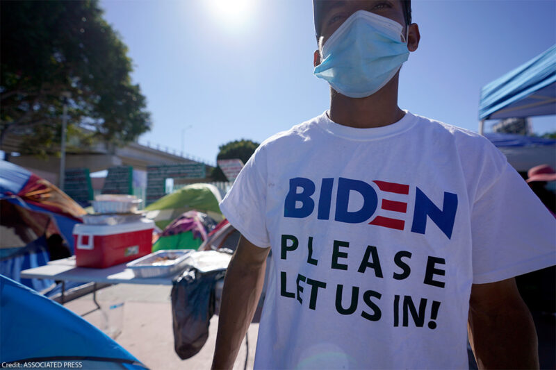 A man seeking asylum in the United States wears a shirt that reads, "BIDEN PLEASE LET US IN!," as he stands among tents that line an entrance to the border crossing in Tijuana, Mexico.