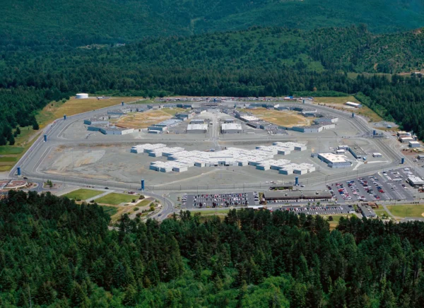 An aerial view of Pelican Bay Prison in Northern California. A large concrete lot with mutliple campuses and scorched green recreational areas is surrounded by a sea of green trees.