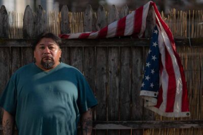 Margarito Casta​​ñon Nava looks into the camera as he stands next to an American flag hanging from a wooden fence behind him on his right.