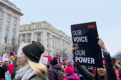 A protestor holding a sign with an ACLU logo that says Our Voice is Our Power.