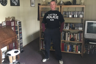 Robert Frese standing in a livingroom wearing a black t-shirt with a message in white lettering reading "EXETER POLICE LIE."