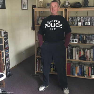 Robert Frese standing in a livingroom wearing a black t-shirt with a message in white lettering reading "EXETER POLICE LIE."