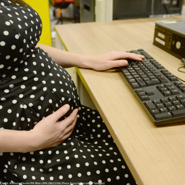 Pregnant person sits at a desk with one hand resting on their stomach and the other on a keyboard.