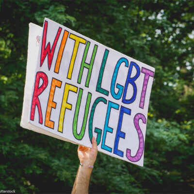 A hand holding up a "With LGBT Refugees" sign.