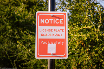 A sign attached to a metal pole with while letters on a red background reading "NOTICE _ LICENSE PLATE READER 24/7 - Flock Safety".