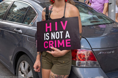 A demonstrator, leaning against a light blue car and with their face hidden holds a sign reading "HIV IS NOT A CRIME".