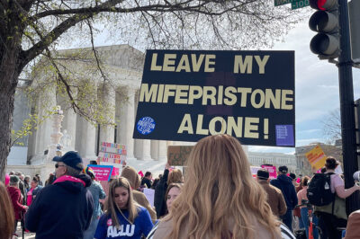a sign saying "leave my mifepristone alone!" is foregrounded with protesters in front of the supreme court.