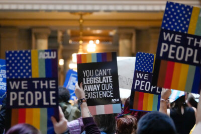 Individuals in a group holding ACLU-branded signs saying "We the People," and "You Can't Legislate Away Our Existence."