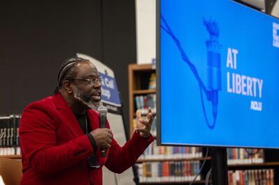 Ian Manuel stands and delivers a poem at the library with bookshelves in the background.