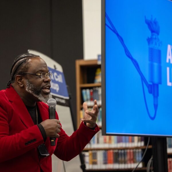 Ian Manuel stands and delivers a poem at the library with bookshelves in the background.