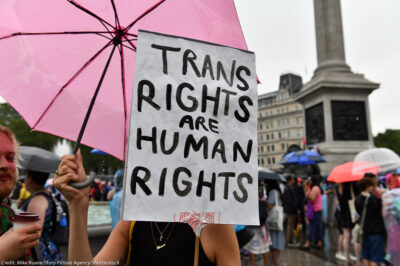 A protestor holds a sign (with a white background and black letters) reading "TRANS RIGHTS ARE HUMAN RIGHTS".