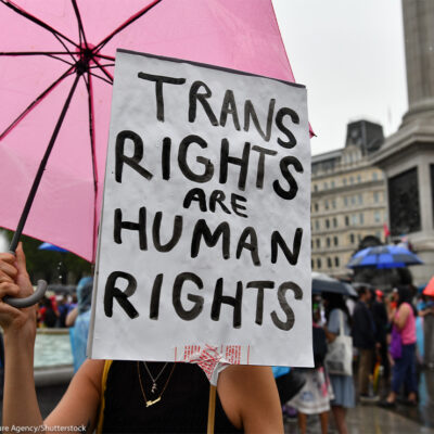 A protestor holds a sign (with a white background and black letters) reading "TRANS RIGHTS ARE HUMAN RIGHTS".