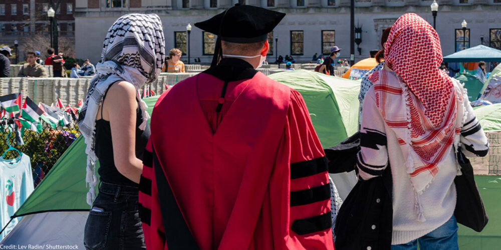 Two individuals wearing keffiyeh scarves standing on either side of an individual wearing graduation garb on a campus.